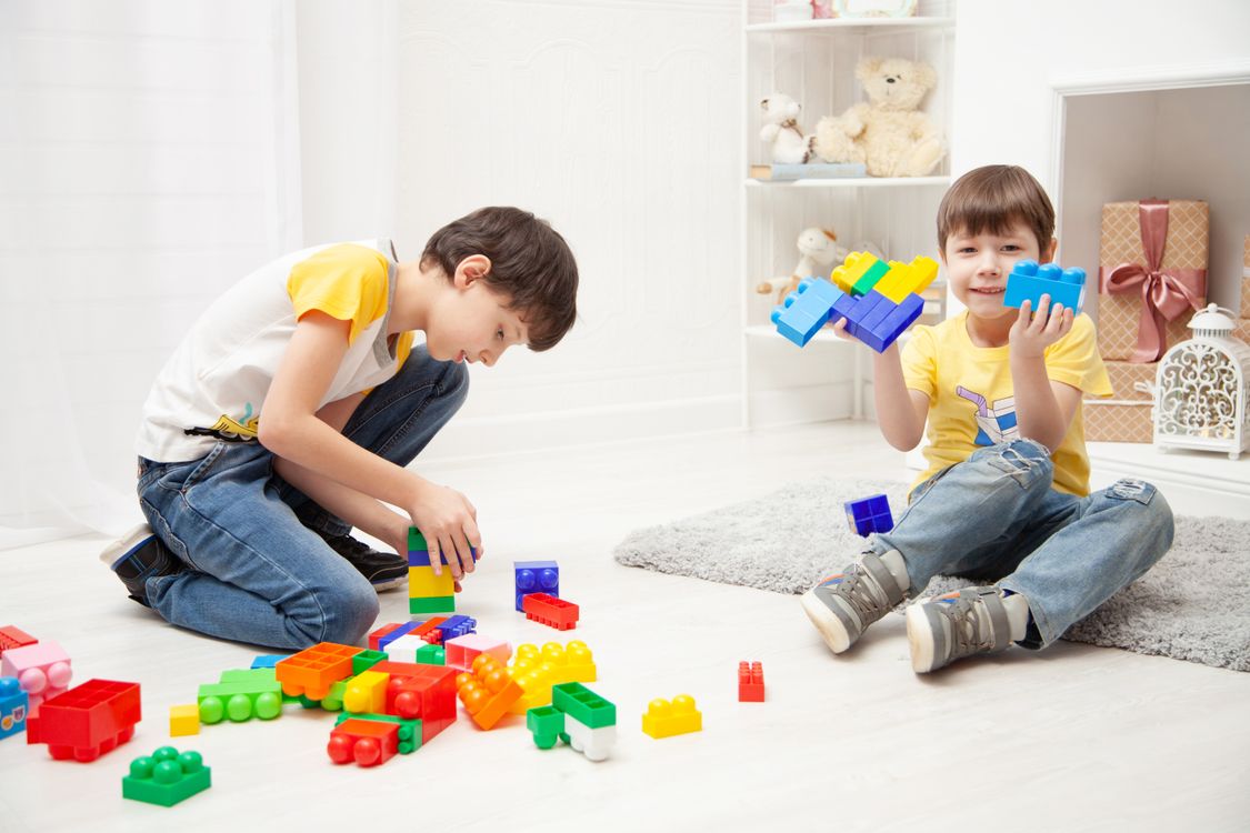 Doi copii construiesc împreună cu piese colorate, pe covor